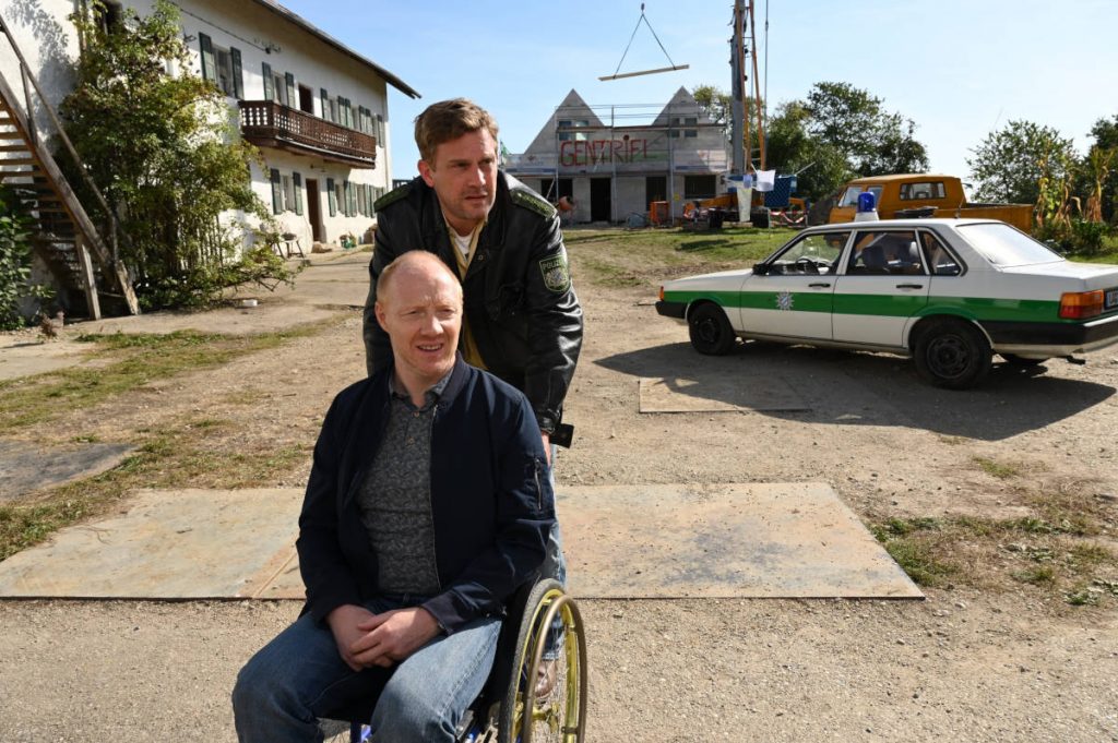 Das Bild zeigt die Ermittler Eberhofer (Sebastian Bezzel) und Birkenberger (Simon Schwarz) zu sehen. Eberhofer schiebt Birkenberger mit dem Rollstuhl.