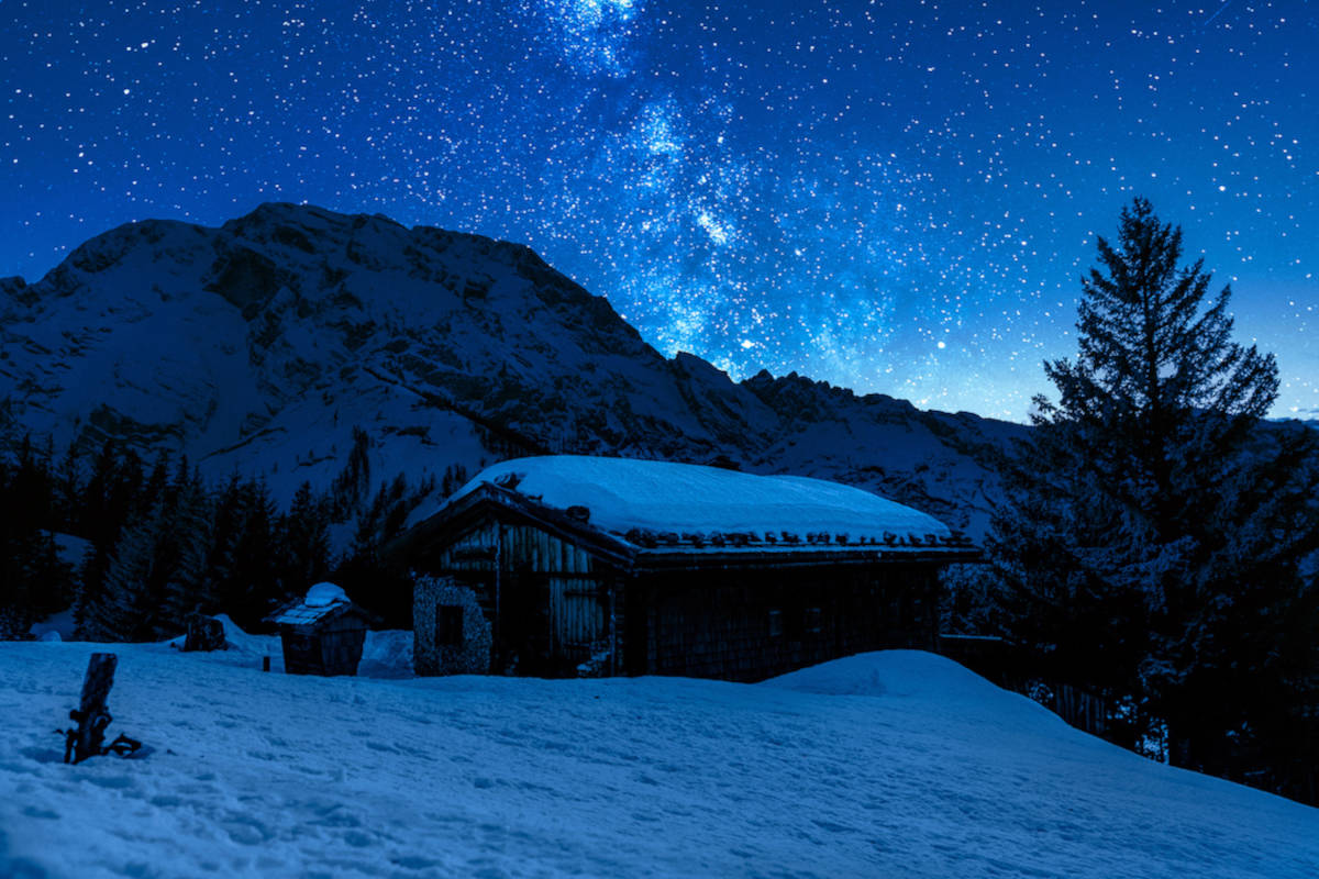 Das Bild zeigt eine kleine Hütte auf einem verschneiten Berg mit Sternenhimmel im Hintergrund.