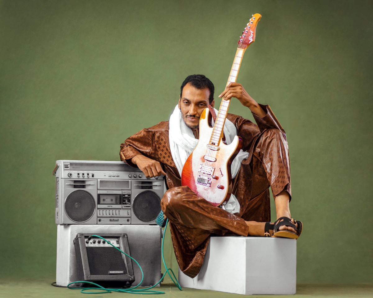Bombino kündigt Deran-Nachfolger-Album an: Das Bild zeigt den Künstler auf einer weißen Box daneben steht ein Ghettoblaster, an dem seine E-Gitarre angeschlossen ist.