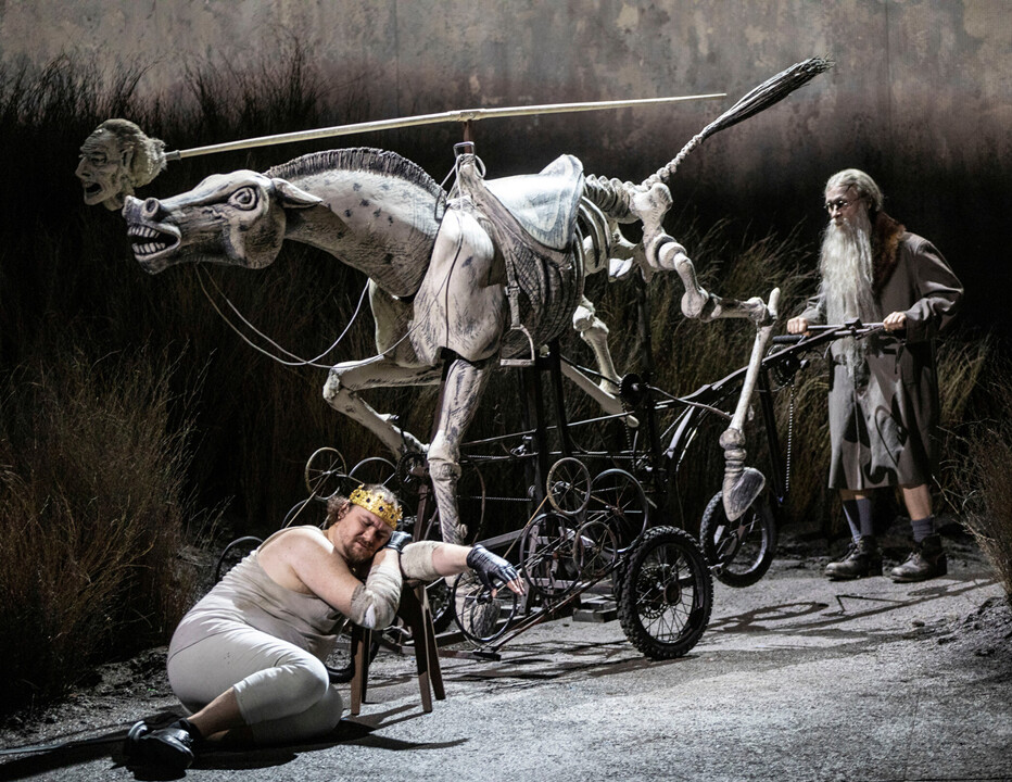 Der Goldene Hahn - Eine bildgewaltige Aufführung in der Komischen Oper Berlin. Das Bild zeigt den König Doden erschöpft auf einem Schemel liegend. Im Hintergrund ein e mystische Pferdegestallt, die von einem Mann mit einem langen Bart geschoben wird.