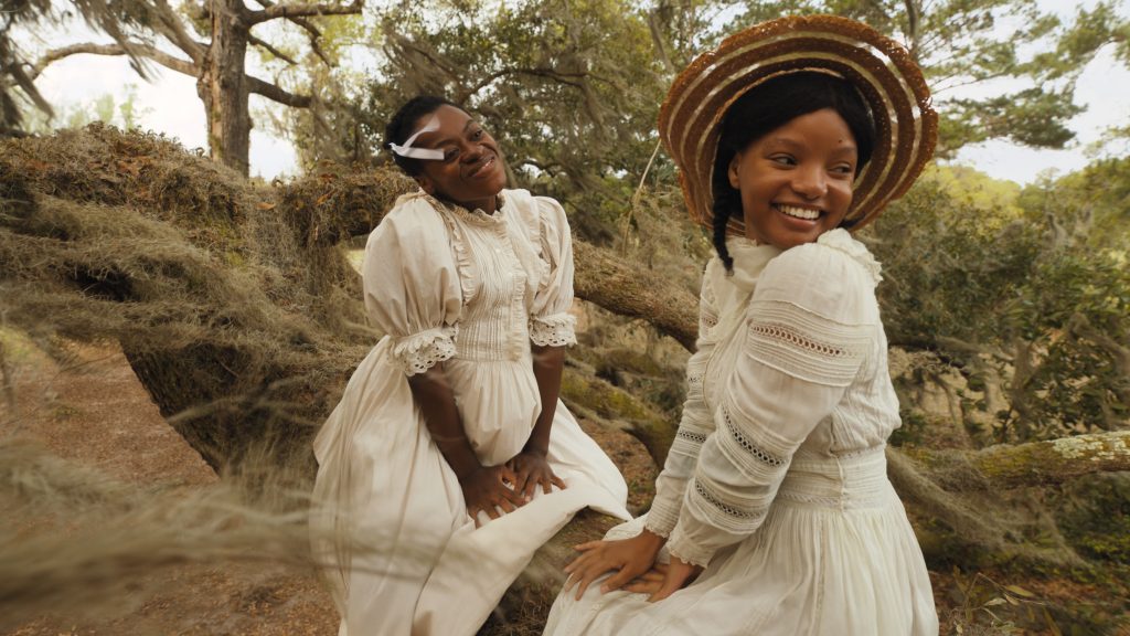 Die Farbe Lila - Die Neuauflage des Klassikers kommt 2024 in die Kinos. Das Bild zeigt zwei schwarze Mädchen in weißen langen Kleidern lachend auf einem Baumstamm sitzen. Eines der Mädchen hat einen Strohhut auf und blickt über die Schulter.