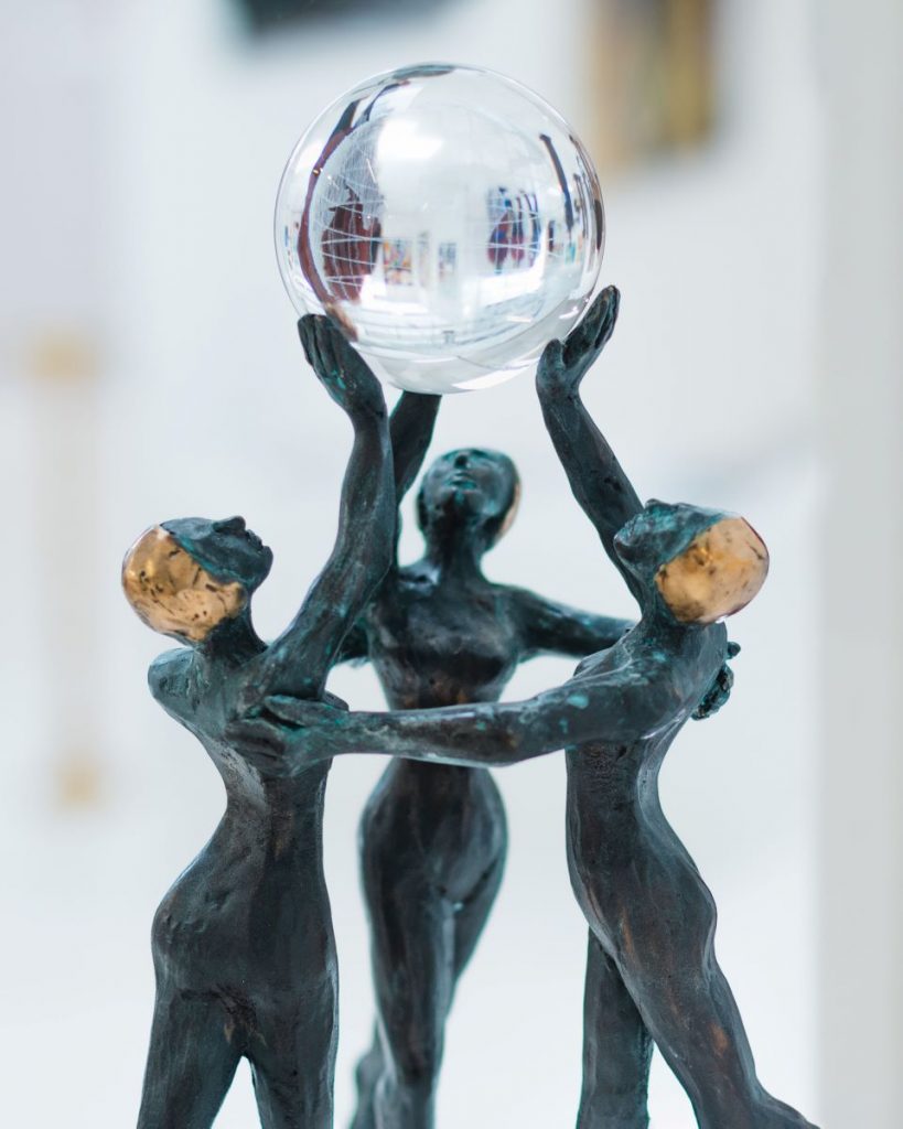 Das Bild zeigt eine schwarze Skulptur: Drei Frauen mit goldenen Haaren bilden einen Kreis und halten eine Glaskugel in die Luft.