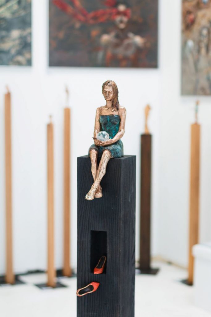 Das Bild zeigt eine Skulptur von einer Frau auf einem schwarzen Sockel. Sie sitzt barfuß, bekleide mit einem grünen schlichten Kleid. In den Händen hält sie eine Kugel. Zu ihren Füßen liegen rote High Heels.