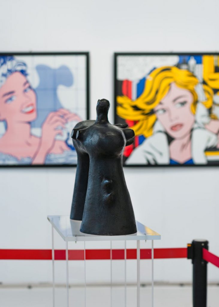 Das Bild von der ARTMUC 2023 zeigt eine schwarze Skulptur auf einem Plexiglassockel. Im Hintergrund sieht man unscharf Pop-Art-Gemälde.