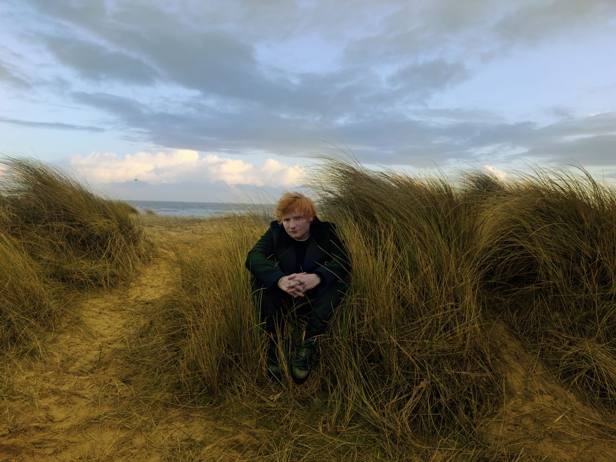 Theresienwiese: Ed Sheeran live. Der Superstar sitzt in den Dünen am Meer. Er ist schwarz gekleidet und sein rotes Haar wird vom Wind verweht.