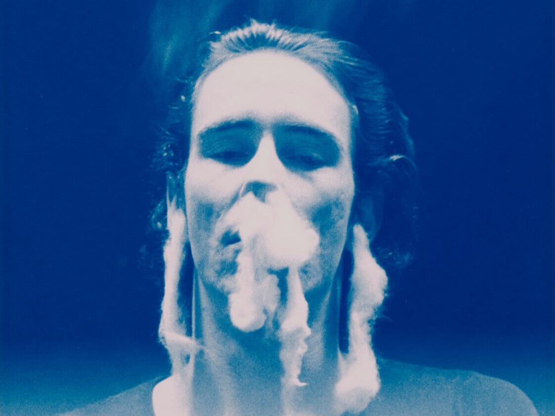 Mike Kelley Kunstausstellung: Ghost and Spirit - Das Bild zeigt den Künstler in einem blau calorisierten Bild, wie er dichten Rauch aus seinen Ohren und Nase bläst. Der Rauch steigt kurioser Weise nach unten.