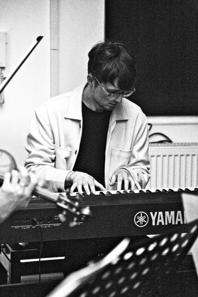 Fabian Kratzer im Studio. Das Bild zeigt den Komponisten an einem Yamaha Keyboard in einer weißen Jacke und schwarzem T-Shirt. Er trägt eine Brille. Auch ist angeschnitten einen Geige und ein Notenständer zu sehen.