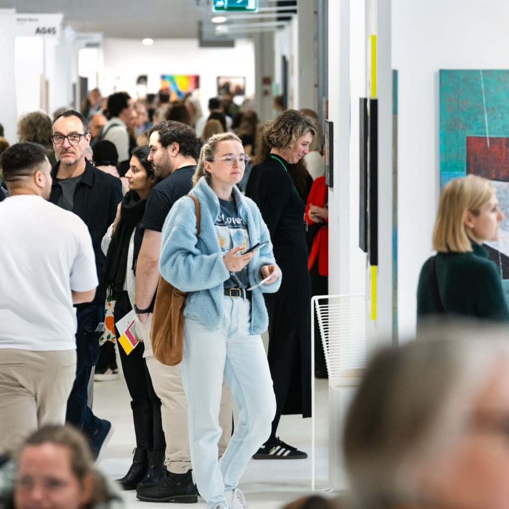 Eine Frau mit blauer Jacke steht im Vordergrund, während viele Menschen durch die Ausstellung der ARTMUC Oktober 2025 gehen.