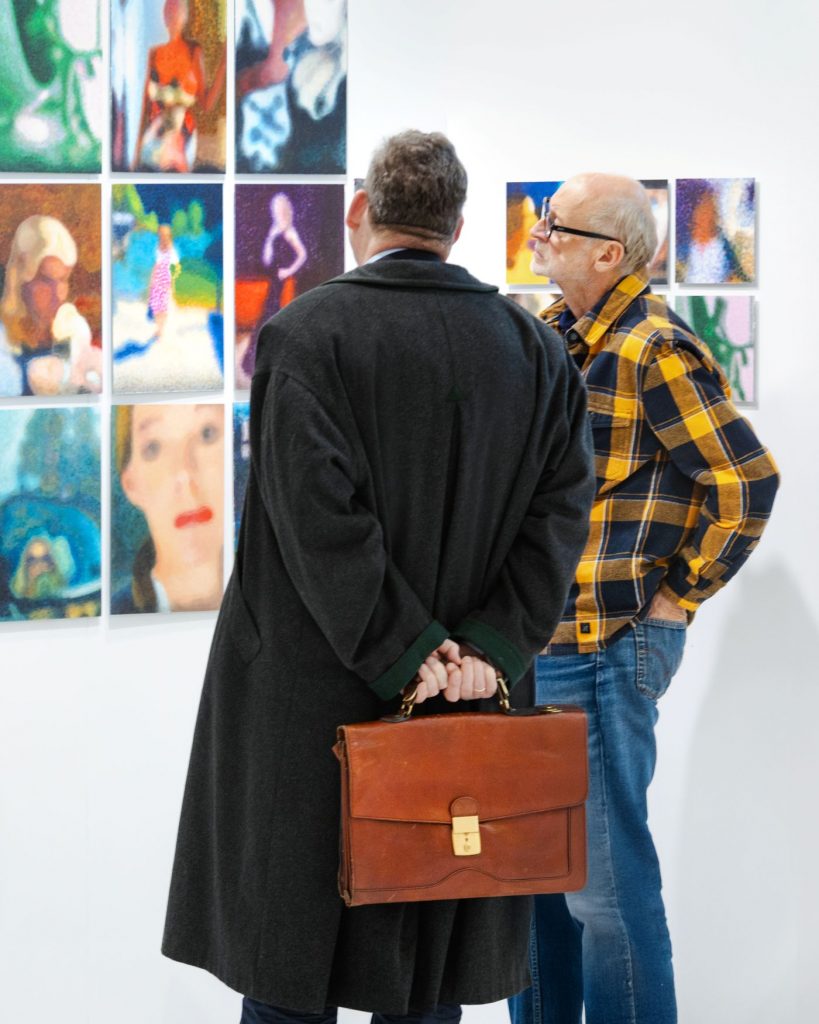 Zwei Männer stehen vor einer Wand mit mehreren bunten Gemälden.