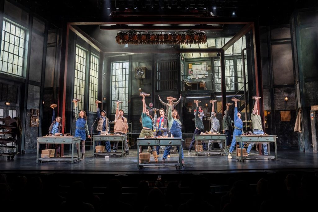 Ensemble auf der Bühne in einer Fabrikszene, Darsteller:innen halten Stiefel hoch – Szene aus Kinky Boots München.